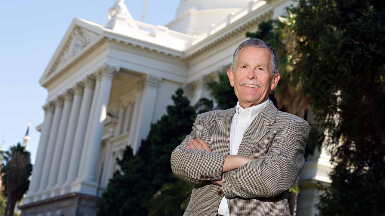 Smiling man in gray blazer, arms crossed, in front of white-columned government building