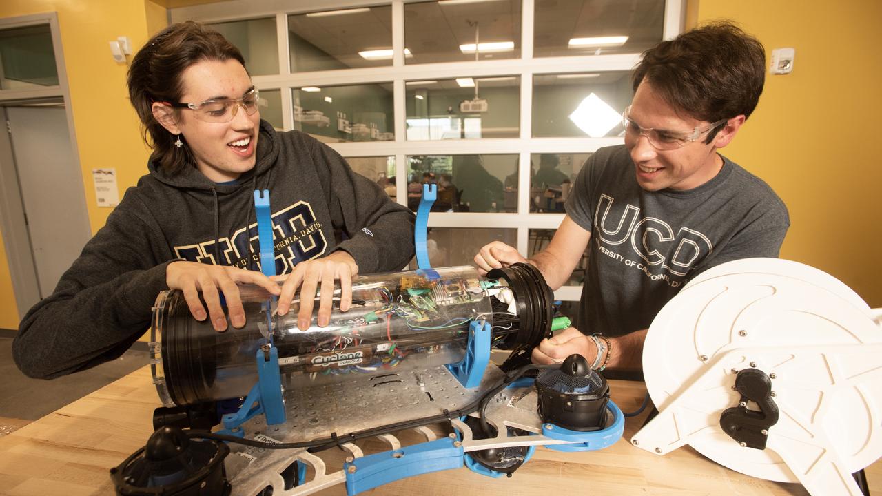 Two students smiling as they work on a blue-accented robot on a table.