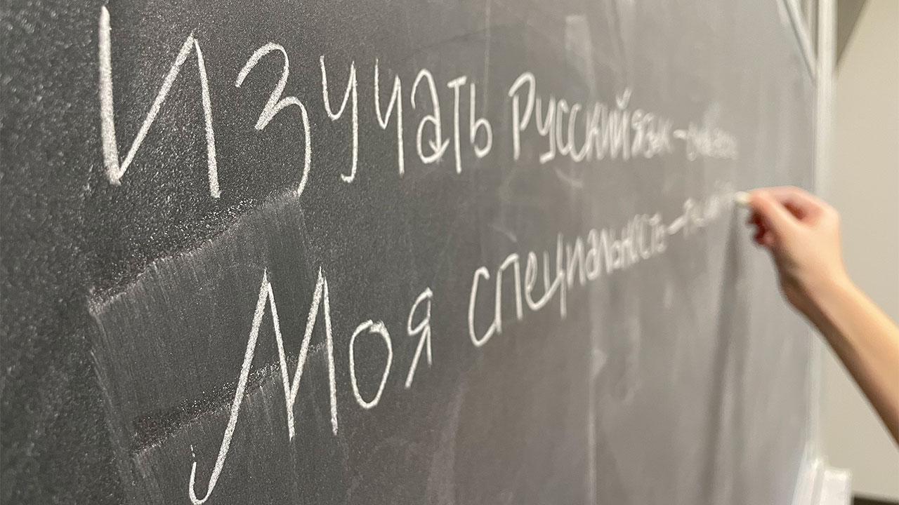 A hand writes on a blackboard in white chalk. The words are Russian, printed in Cyrillic.