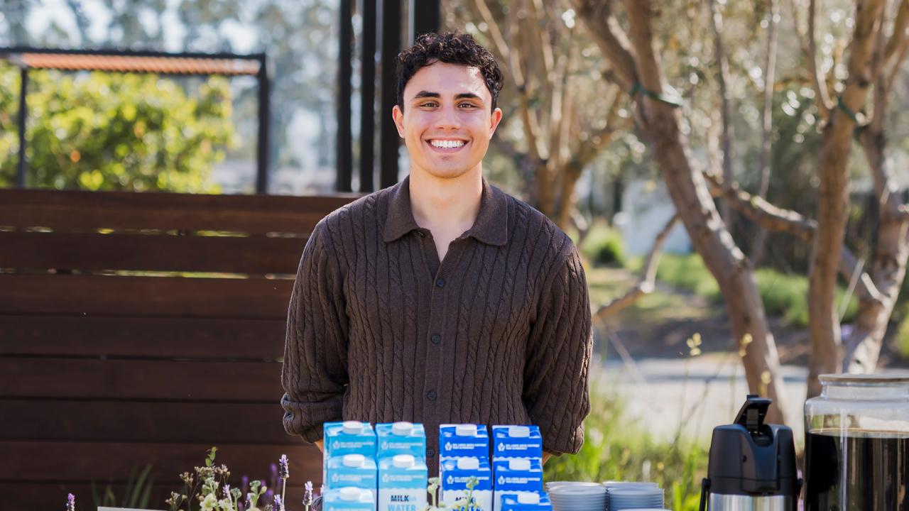 Smiling man outdoors behind table of blue-and-white milk cartons and coffee maker