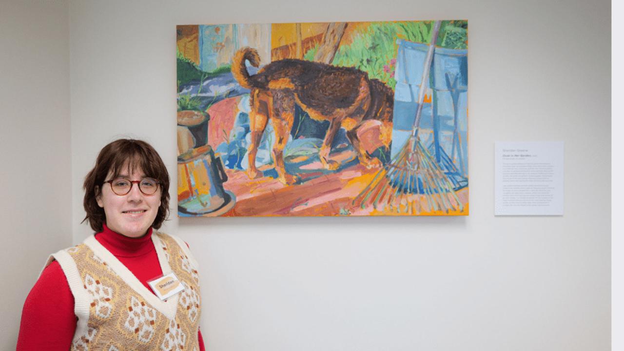A woman in a red shirt and beige vest stands beside a colorful dog painting in an art gallery.