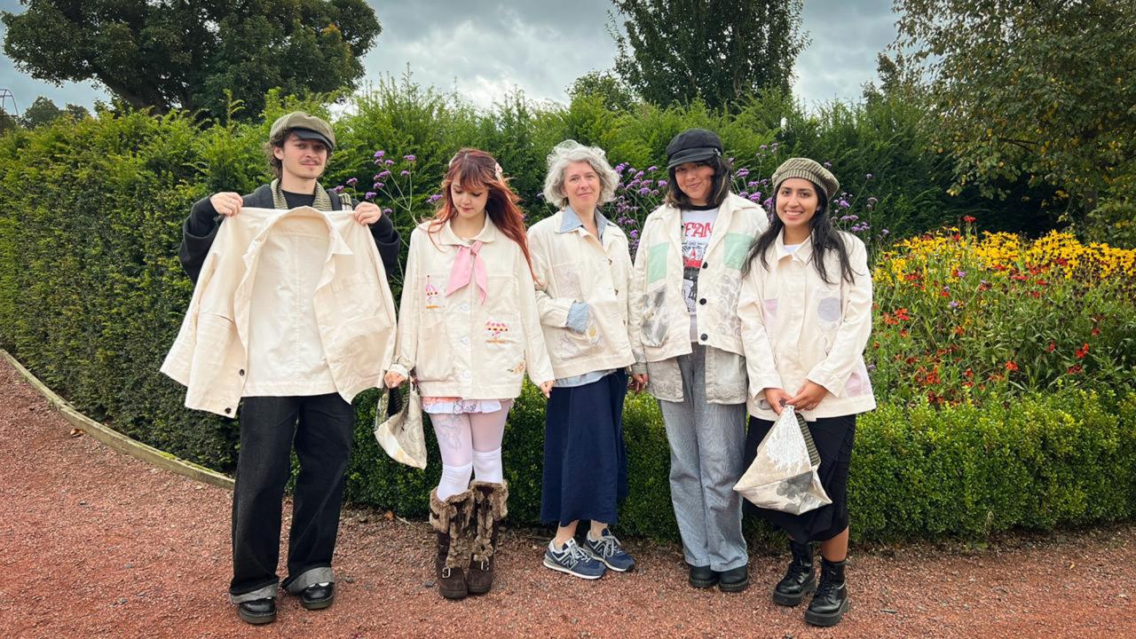 Five people wearing light-colored jackets stand in a garden with colorful flowers.