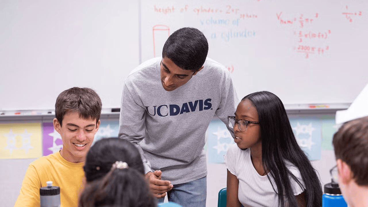 Student wearing UC Davis shirt leans over to speak to younger students in classroom setting