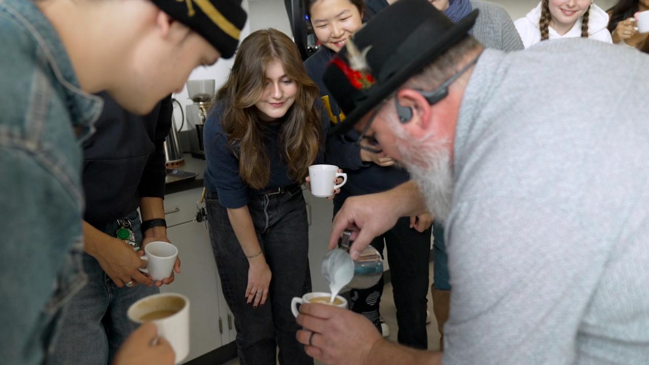 Students gather and watch with excitement as a white-bearded man in a light gray shirt, glasses, black open-ear Bluetooth headphones and a feathered black fedora pours foam into a small, white coffee mug.