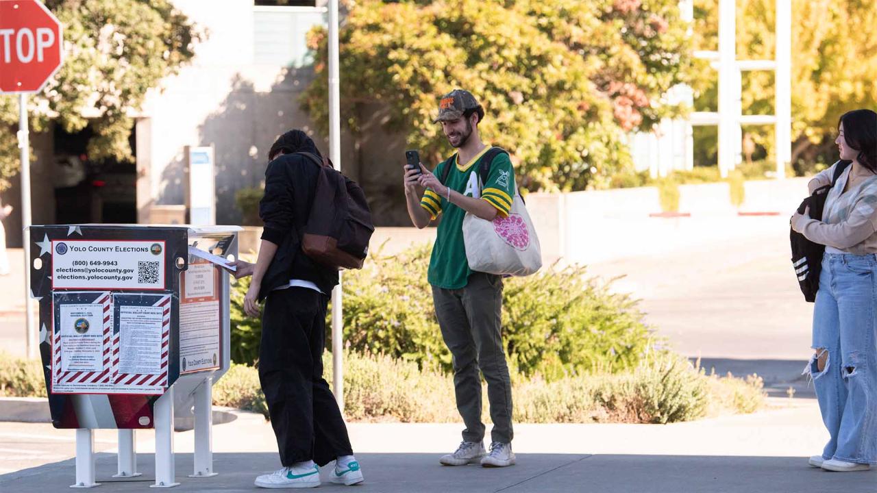 One person drops their ballot into a metal drop box while another takes a photo. A third person waits to drop off their ballot.
