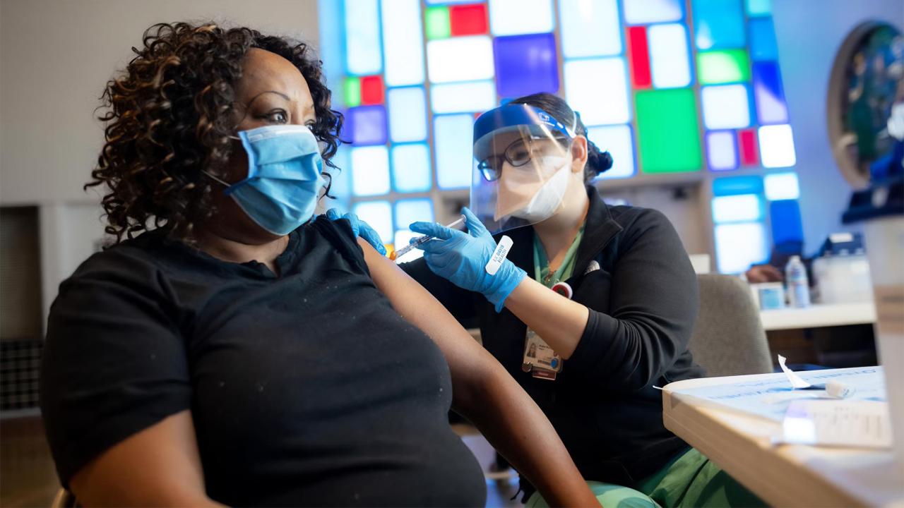 A healthcare worker in protective gear administers a COVID-19 vaccine to a masked individual wearing a black shirt, with a stained-glass window in the background.