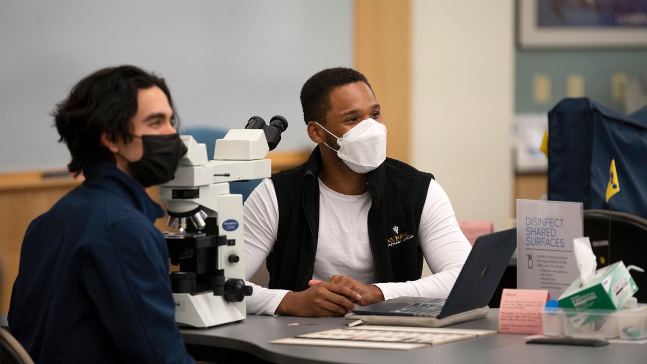 Two students wearing face coverings sit at table in laboratory.