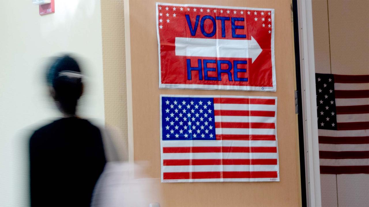Blurred person walks past red 'VOTE HERE' sign and U.S. flag at polling place