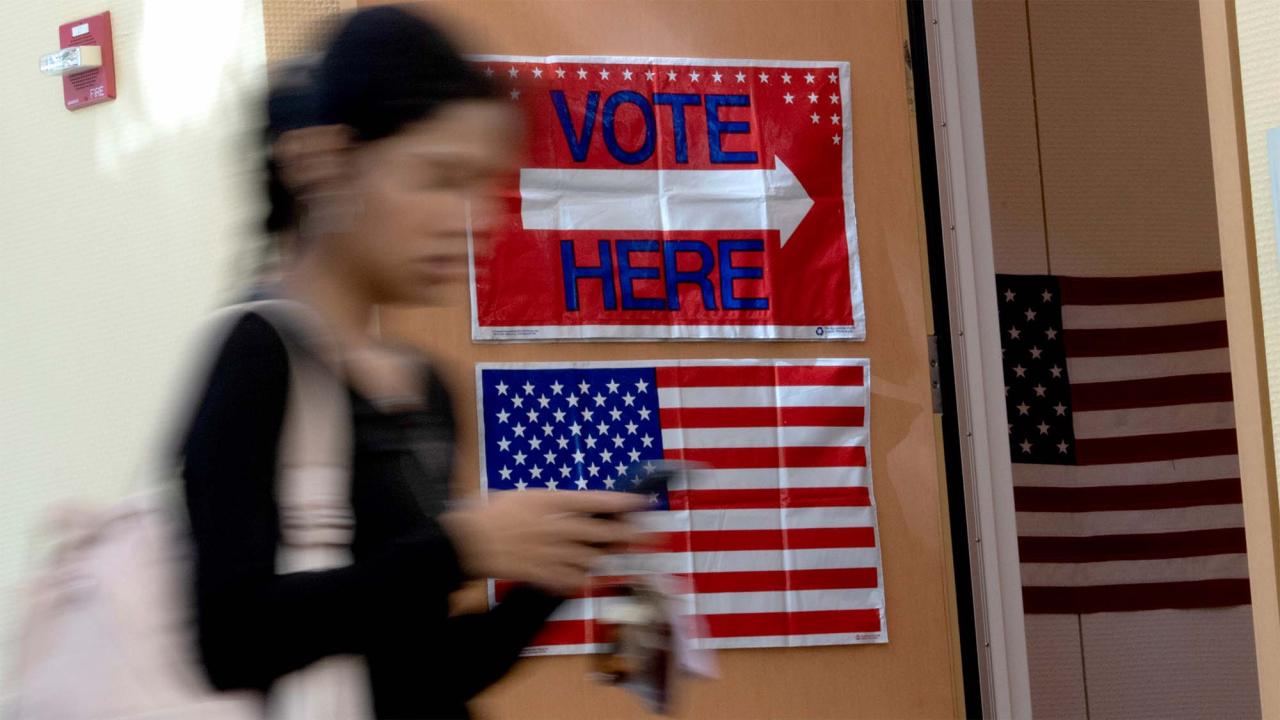 Blurred voter walking past a 'VOTE HERE' sign and American flag in polling place