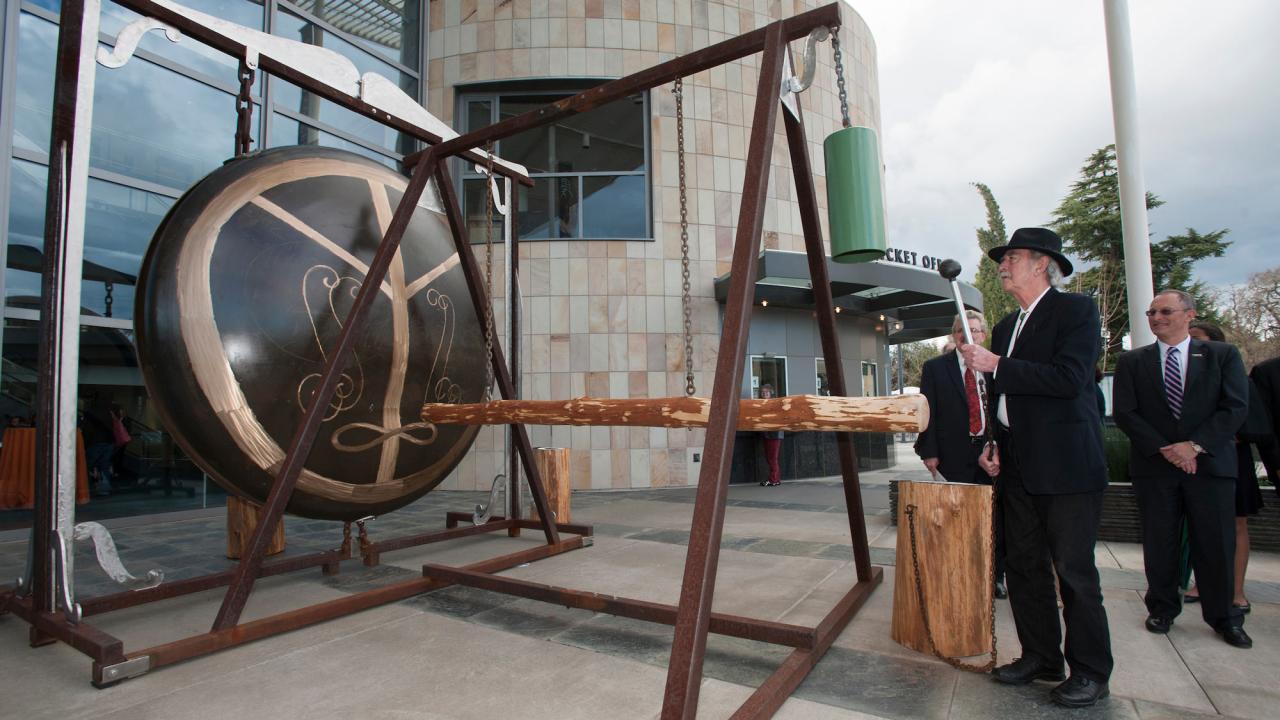 Man holds gong striker, next to Gong sculpture