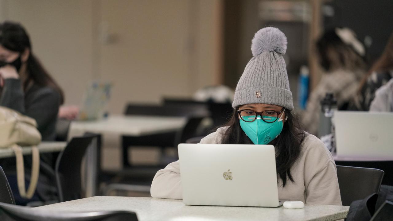 Wiman in mask and knitted cap, at laptop, in Memorial Union