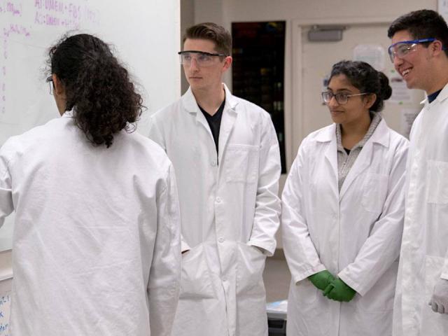 Students in lab coats stand in front of a white board.