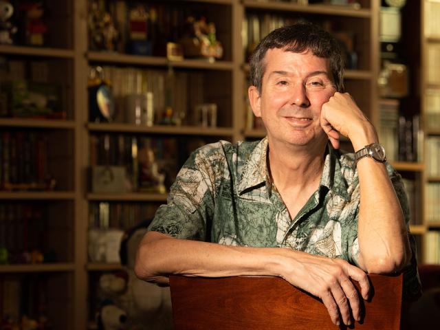 Smiling man in patterned shirt leaning on a chair in warm-lit book-filled room