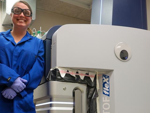 Smiling lab worker in blue coat beside machine decorated with googly eye and paper teeth, photo