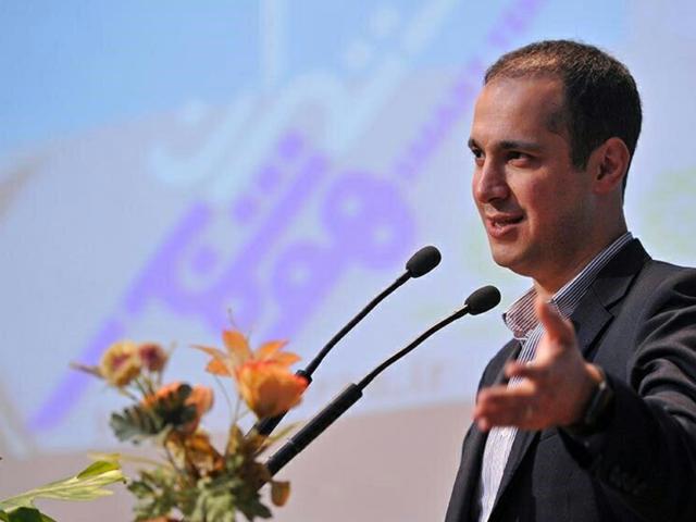 Man in suit speaking at podium with microphones, gesturing near a floral arrangement