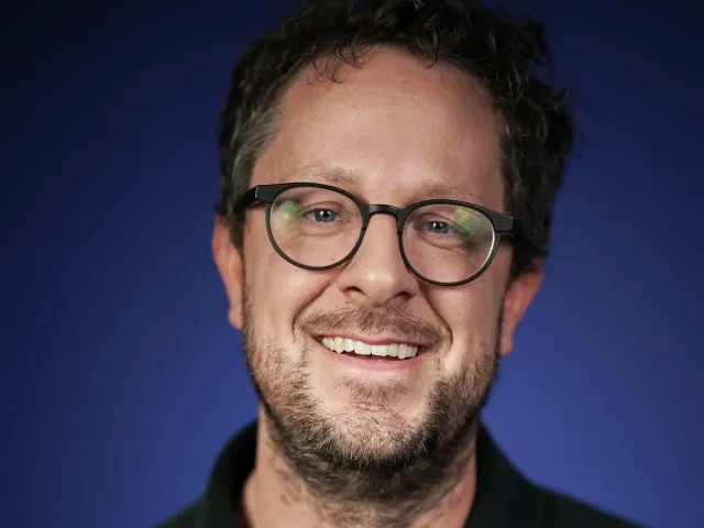 Smiling person in glasses, scruffy beard, black shirt — studio headshot, blue gradient background