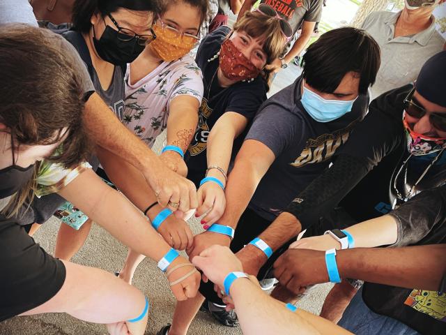 A group of people reach their hands into the center of a the circle and show off blue wristbands