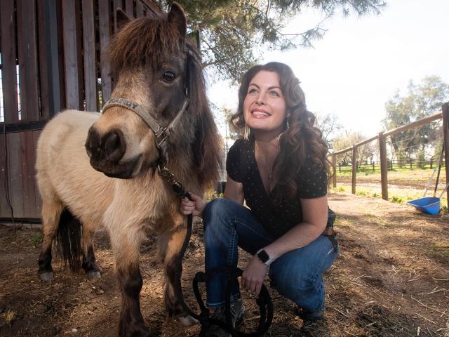 Woman crouching and smiling beside a small horse at a sunny farm