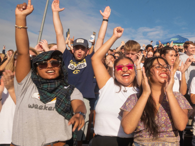 Excited crowd of students cheering at an outdoor event, with several people raising their arms and smiling in the sunlight.