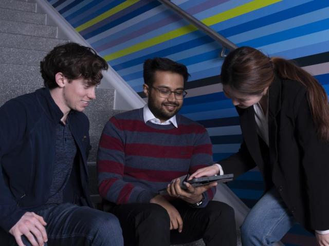 three students sitting on steps talking and looking at a device