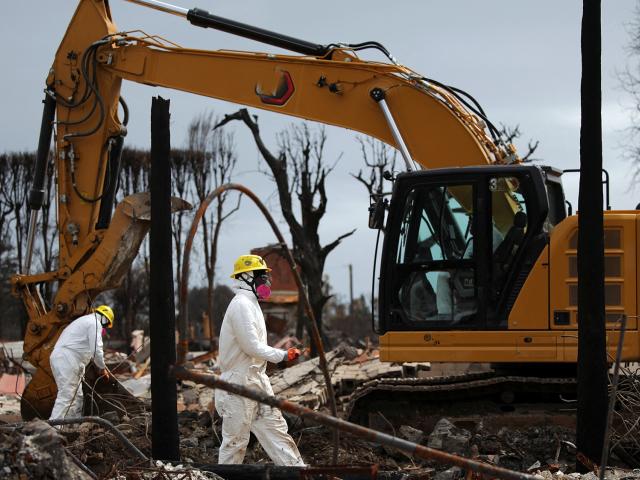 Two workers in white hazmat suits beside a yellow excavator at a muddy, bare-treed site