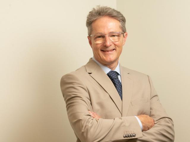 Smiling man in beige suit and blue tie, arms crossed against neutral background