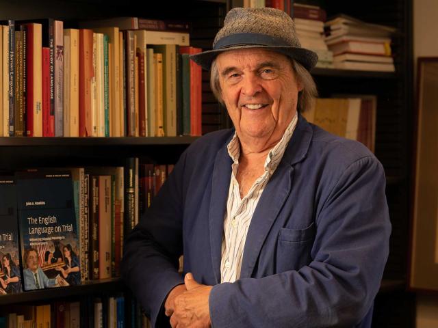 Smiling man in hat and blue jacket by bookshelf, warm-lit photograph.