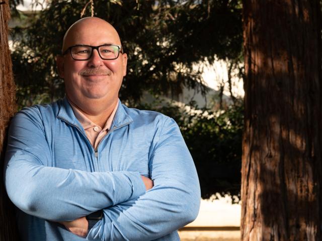 Smiling man with glasses stands confidently against a tree, arms crossed.