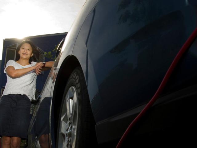 A woman poses next to a car that is plugged in to an electric charging station