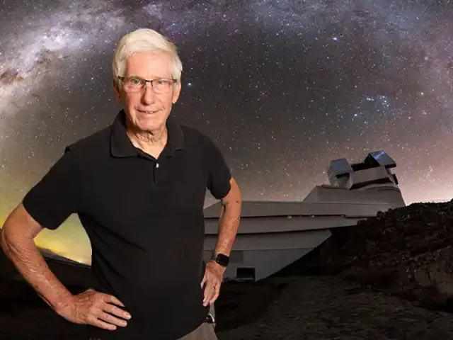 Older man with white hair and glasses in black shirt standing before observatory under Milky Way