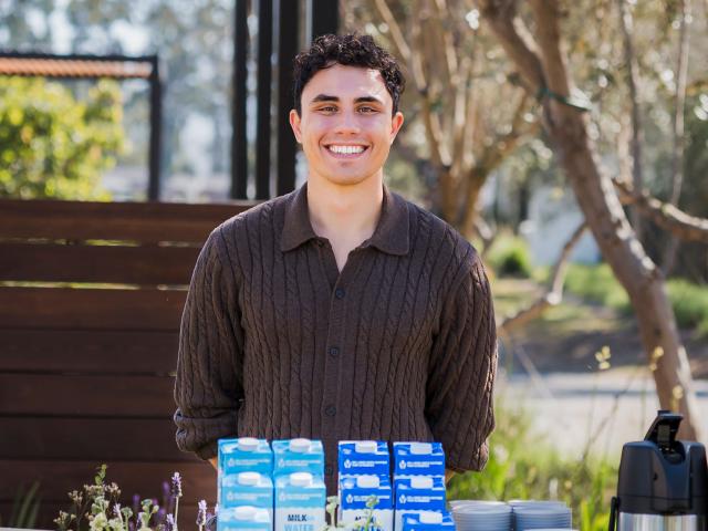 Smiling man outdoors behind table of blue-and-white milk cartons and coffee maker