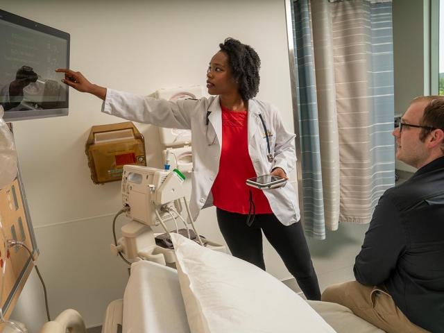 Scene in a hospital room with a woman nurse or doctor in a white coat and stethoscope pointing to a computer screen mounted on the wall displaying data. A man, presumably a patient, sits on the end of the hospital bed. 