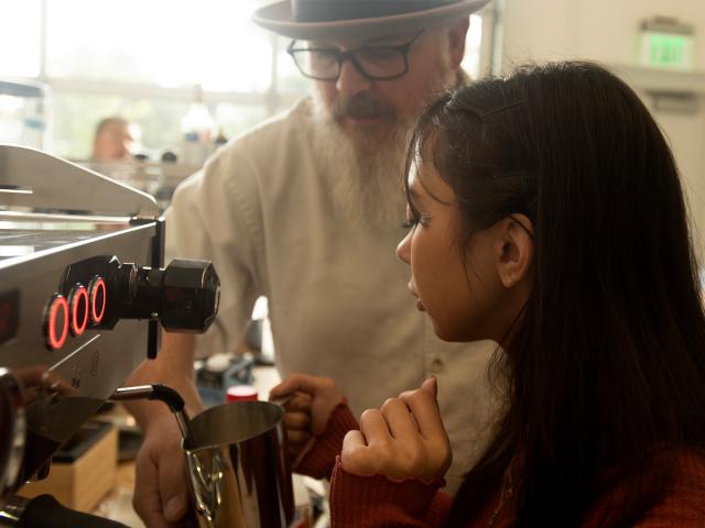 A student in a burgundy long-sleeve shirt carefully uses the milk frother on an espresso machine while being supervised by a man with a white beard, wearing square-rimmed glasses and a fedora.