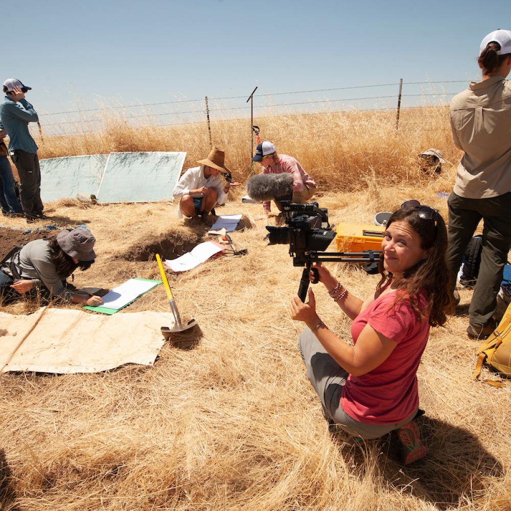 Woman in red shirt with video camera smiles while soil science students sample soil in dry straw landscape