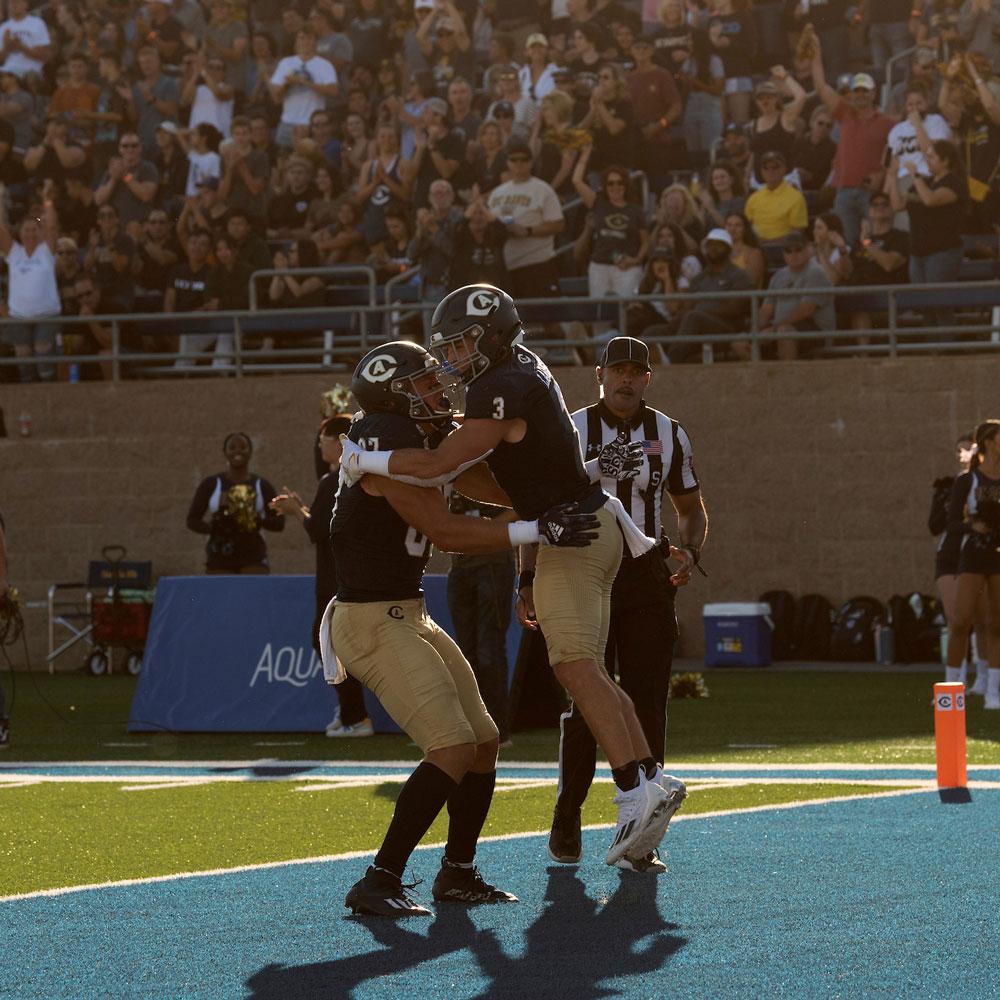 Two college football players in black jerseys hugging on blue endzone turf, crowd behind.