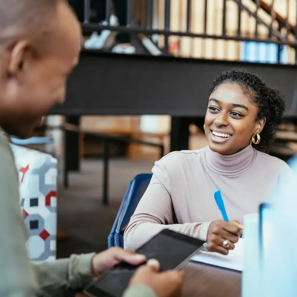 Smiling woman in turtleneck, writing in a notebook while talking with a man across the table
