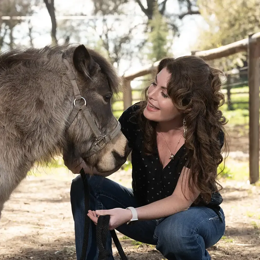 Smiling woman kneels and pets a small donkey in a sunlit paddock, photo