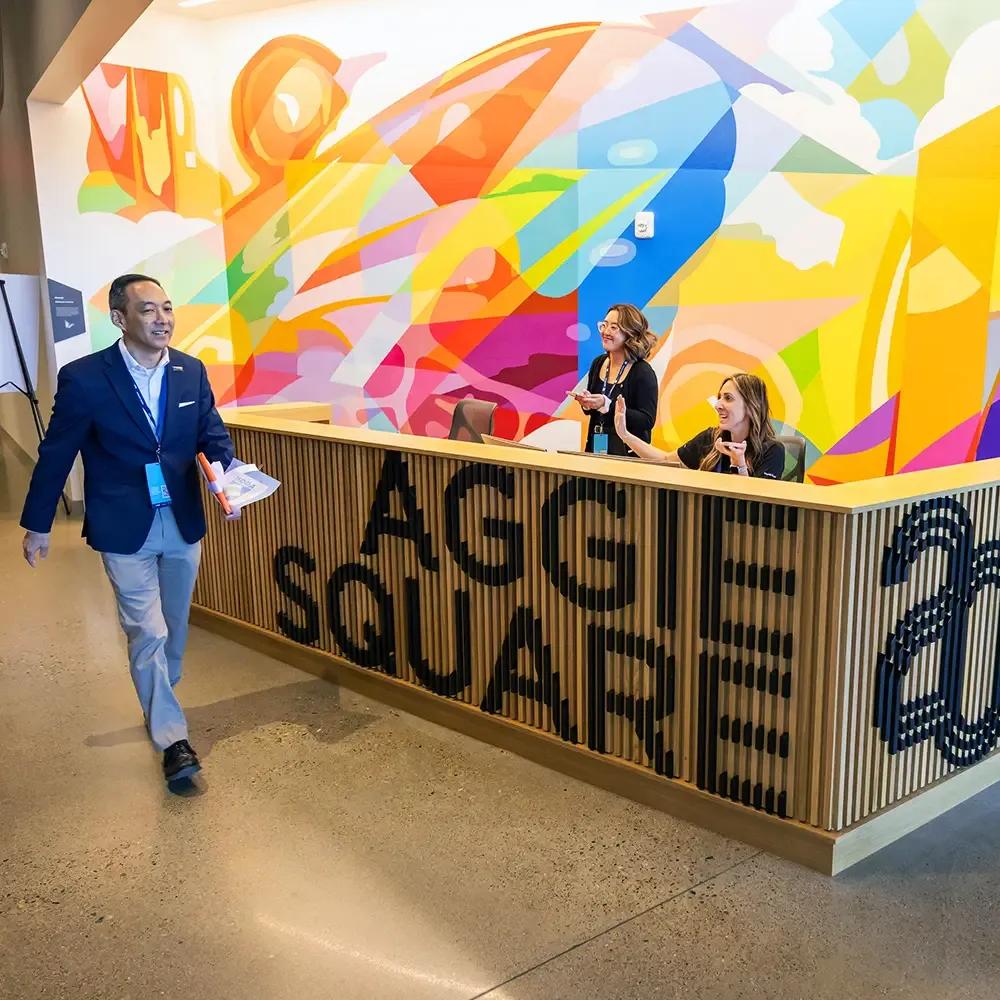 A person walking by a reception desk with Aggie Square written on the side and two employees sitting behind it.