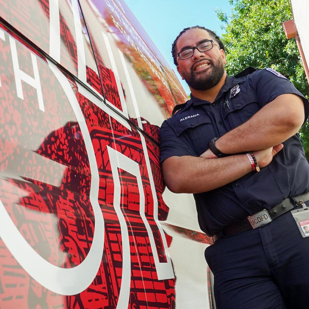 Smiling man in dark uniform, arms crossed, leaning against a red ambulance under blue sky