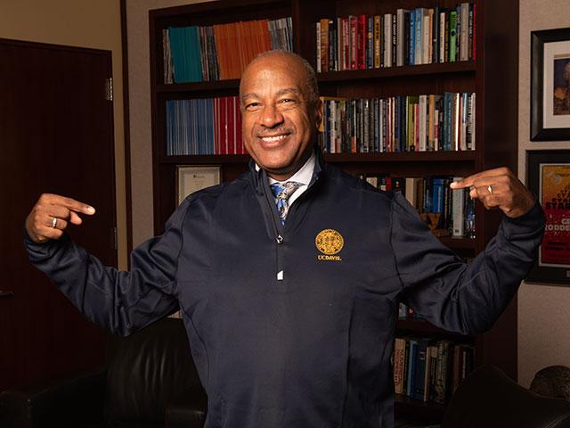 Smiling older man in navy jacket pointing at embroidered logo, bookshelf office background