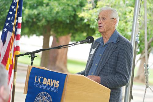 An older man with grey hair and glasses, wearing a blue shirt and jacket without necktie stands at a podium outdoors. A banner on the podium says UC Davis. To the left of the image is a US flag.