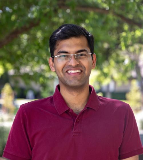 Head and shoulders of a smiling young man with short dark hair wearing a red open necked polo shirt and glasses.