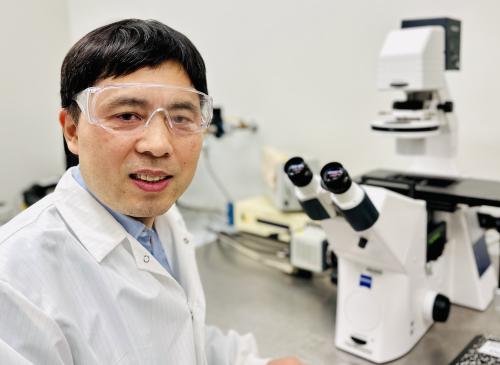 A man in a white lab coat and safety glasses with a microscope in the background.