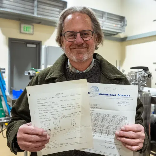 A man with glasses smiles while holding two documents in a lab setting.