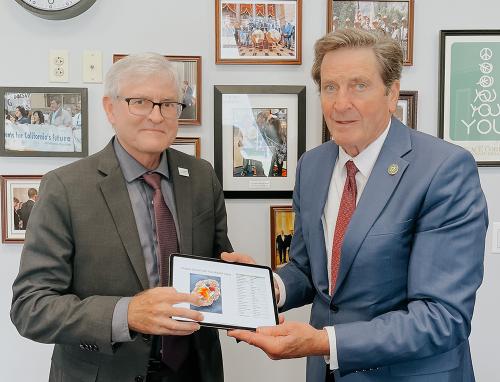 Left, white man in brown suite with white hair and glasses, on right taller white man with brown hair, blue suite and red tie. 