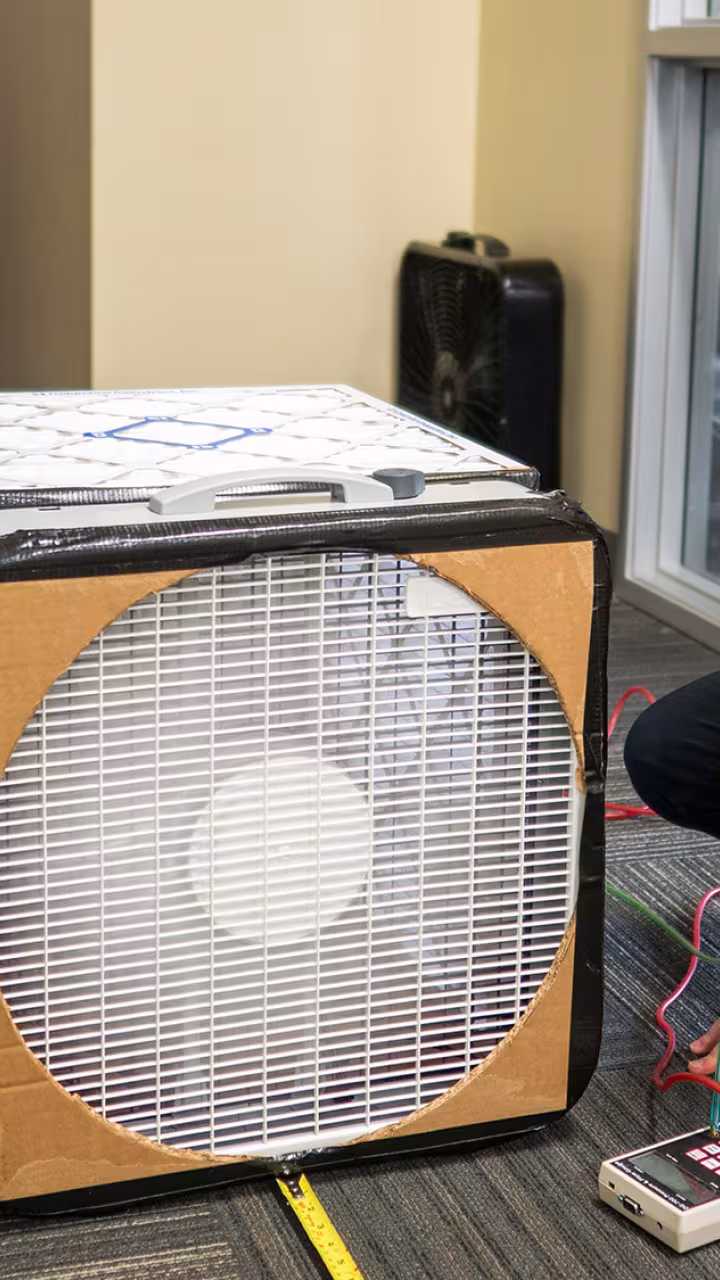 A woman kneels next to a box with air filters on it to monitor the air quality of a room