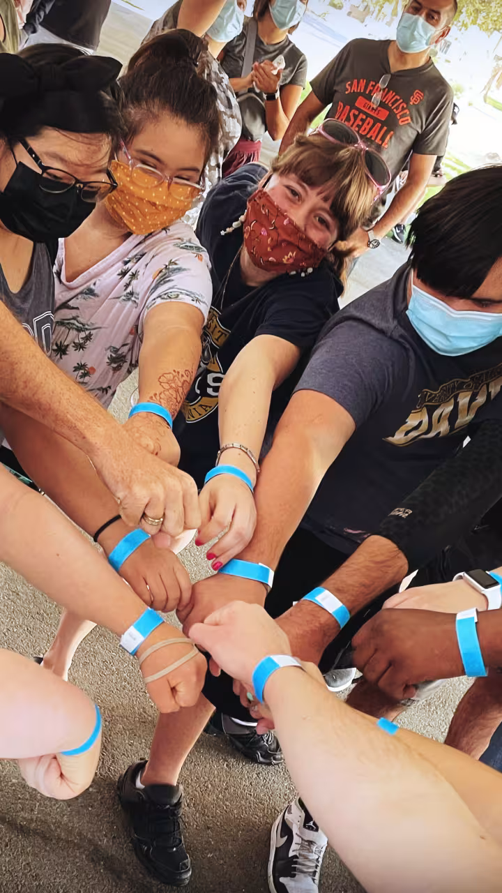 A group of people reach their hands into the center of a the circle and show off blue wristbands