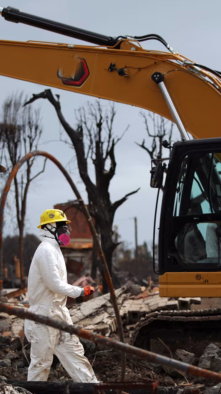 Two workers in white hazmat suits beside a yellow excavator at a muddy, bare-treed site