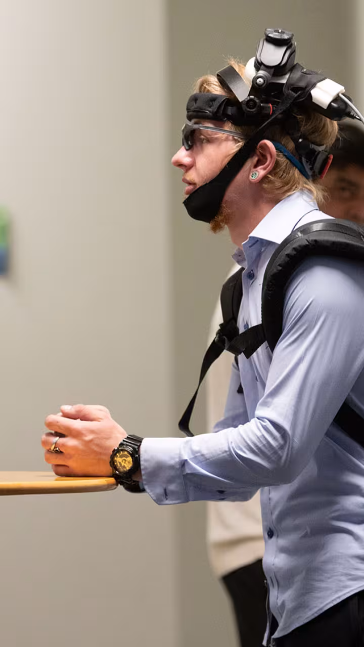 Person wearing head-mounted tracking gear and backpack, leaning on a table in a hallway.