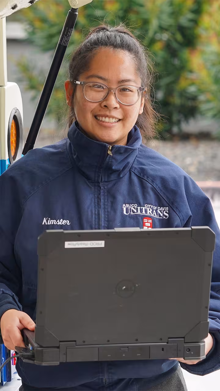 A woman holds a computer outside a bus's rear panel that is open